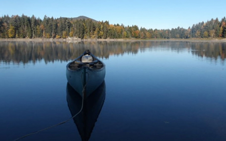 An empty canoe rests on very calm water, reflecting the blue sky and tree-lined shore in the background. 